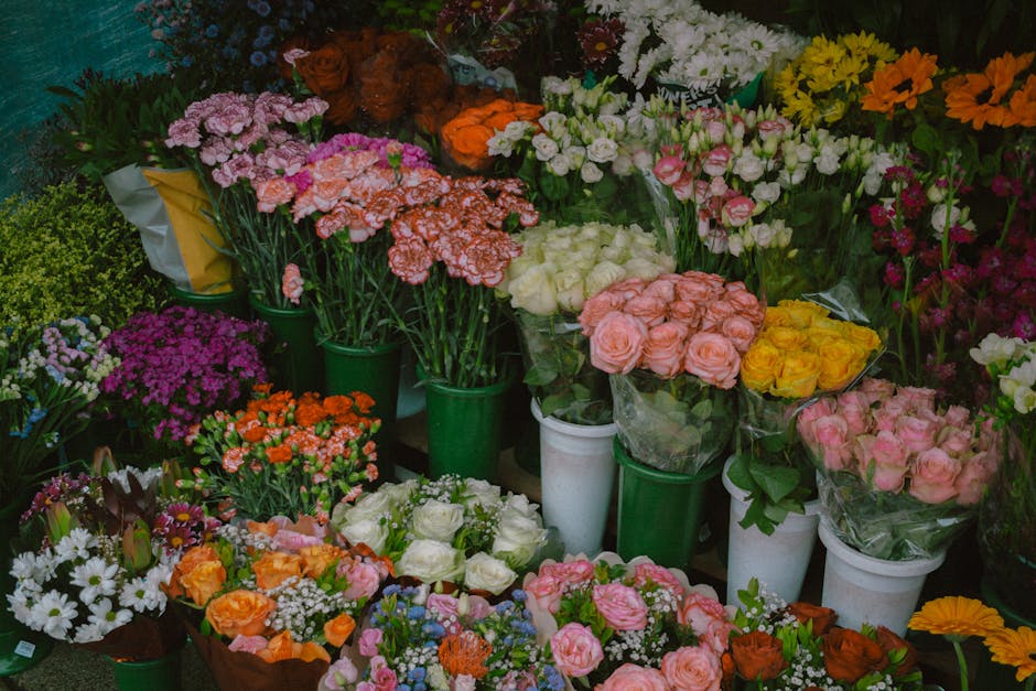 A vibrant display of various fresh flowers arranged in green and white buckets and vases, including pink and peach roses, white roses, orange and yellow tulips, purple asters, daisies, and other color